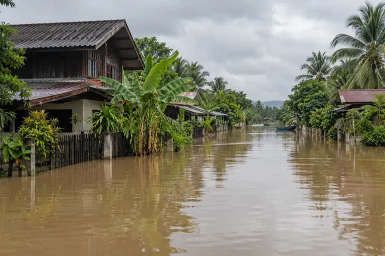 ถนนและบ้านเรือนถูกน้ำท่วมสีน้ำตาลขุ่นในพื้นที่ชนบทไทย ท่ามกลางฟ้าครึ้ม
